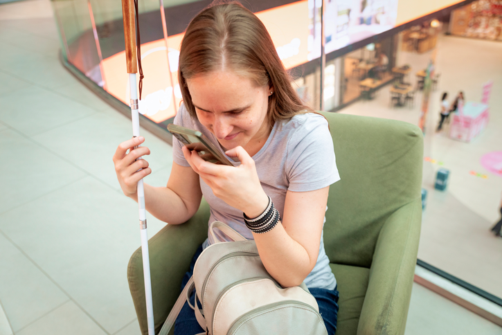 A woman with a white cane holds her smartphone close to her face while sitting in a shopping mall.
