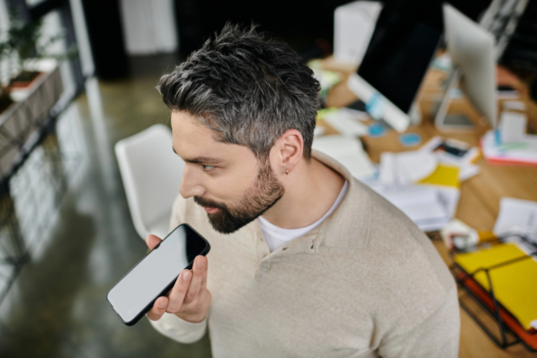 A man speaks into his smartphone while standing in a busy office.
