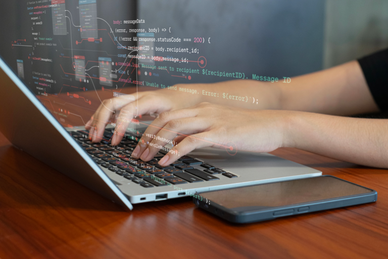 Hands typing on a laptop keyboard with lines of code overlaid on the screen, next to a smartphone on a desk.