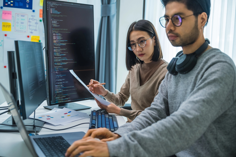 Two developers reviewing code on a monitor against printed notes, with requirements on sticky notes behind them, illustrating a requirements ledger