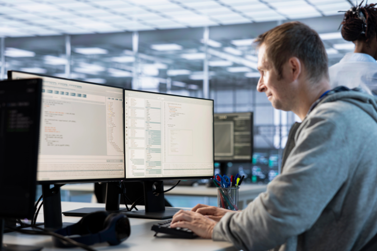 Software developer reviewing code side by side on two monitors in a bright office.