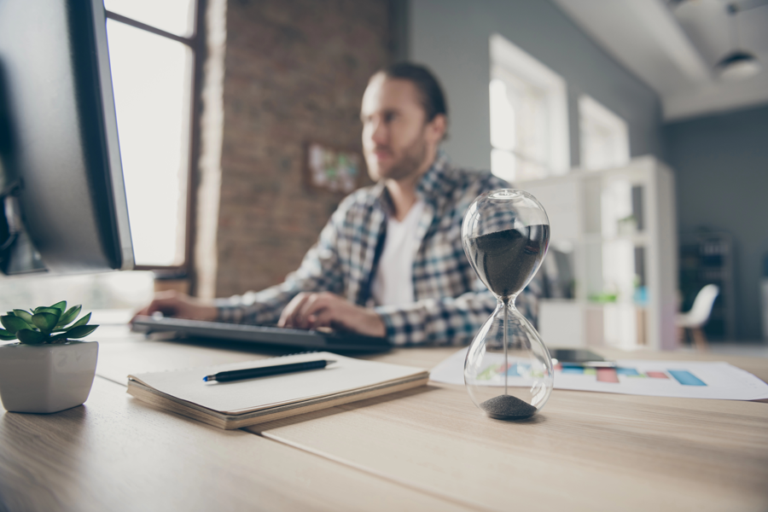 Hourglass with sand narrowing through its neck on a developer's desk, illustrating the AI testing bottleneck.